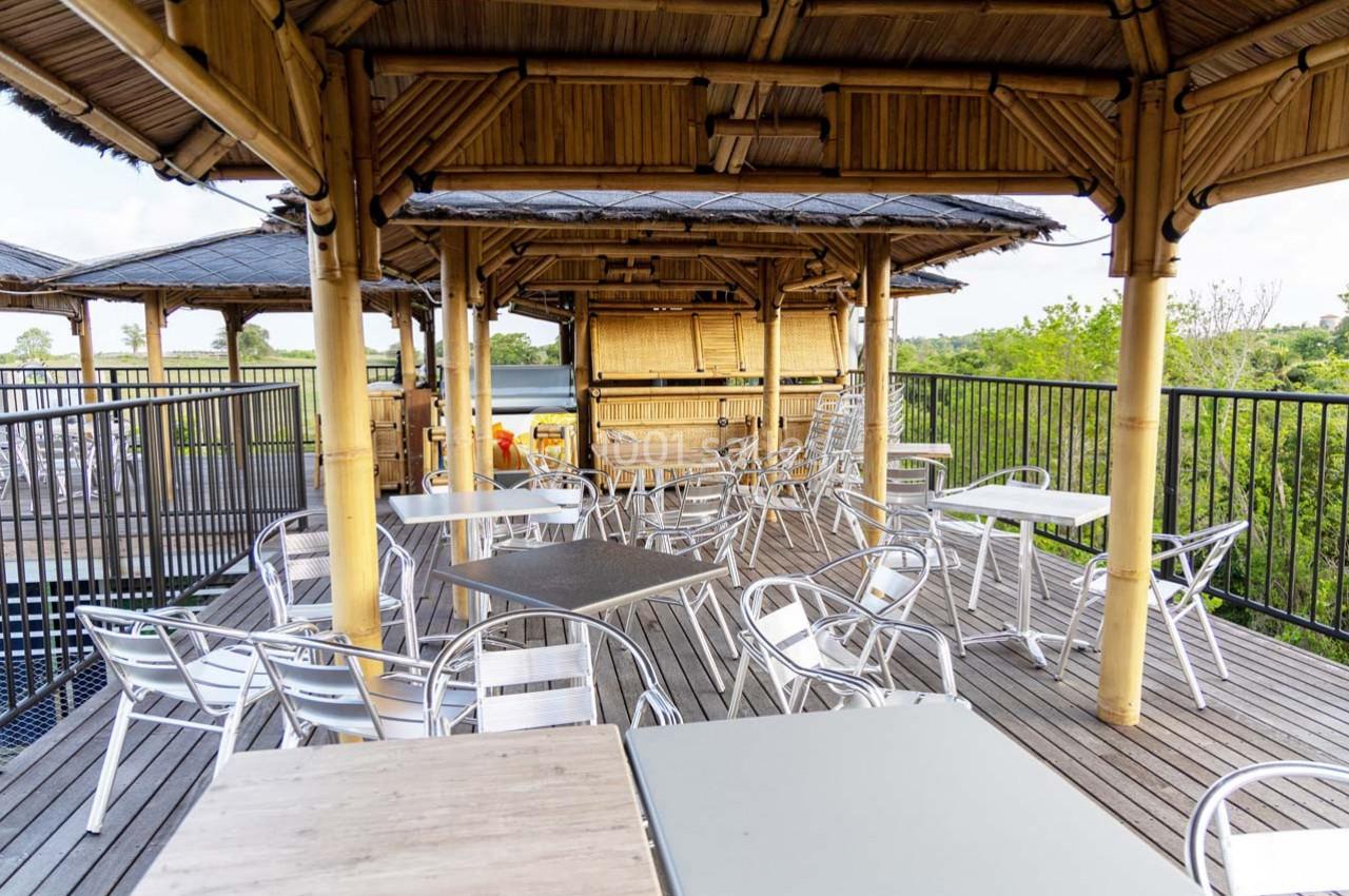 Terrasse en bois avec des tables et chaises en métal sous une structure en bambou, entourée de verdure.