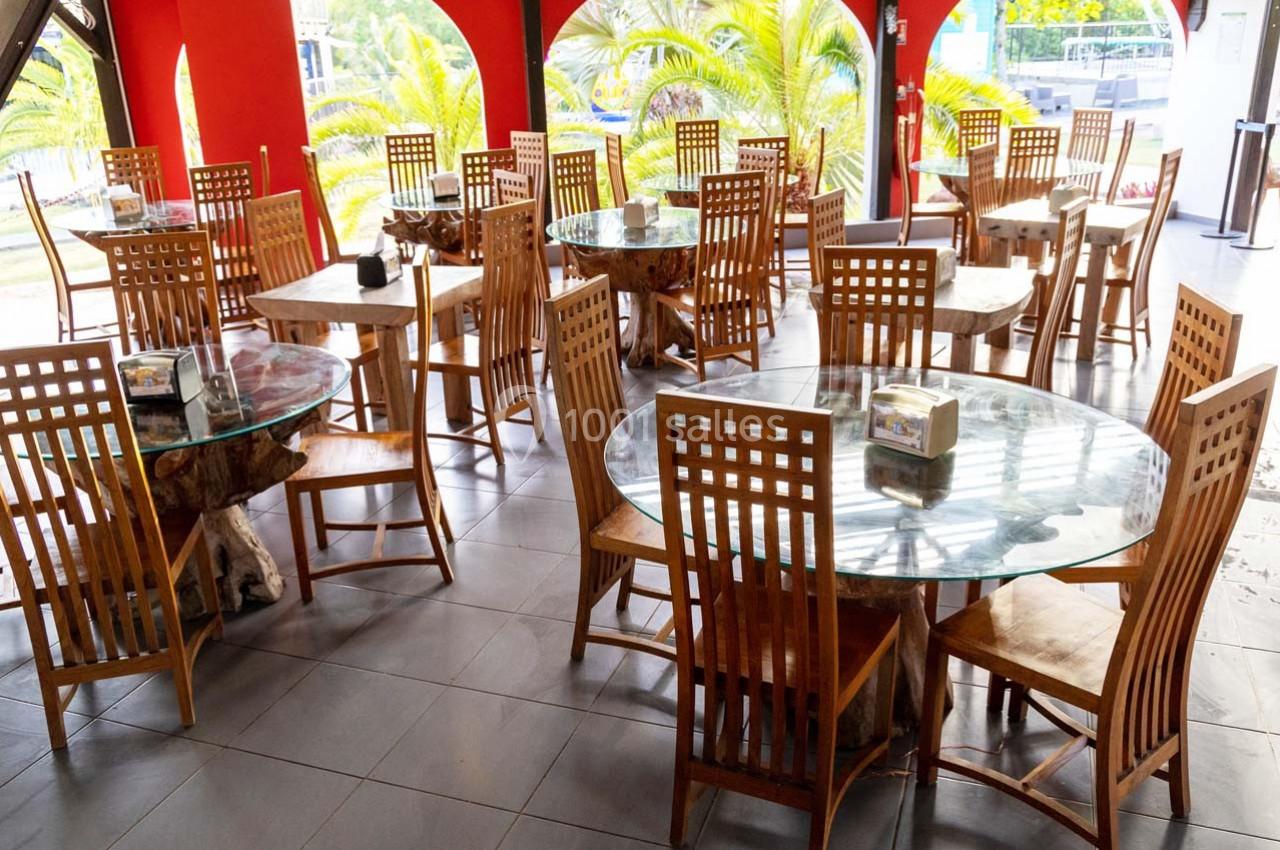 Salle de restaurant avec des tables rondes en verre et des chaises en bois, entourée de grandes ouvertures sur l'extérieur.