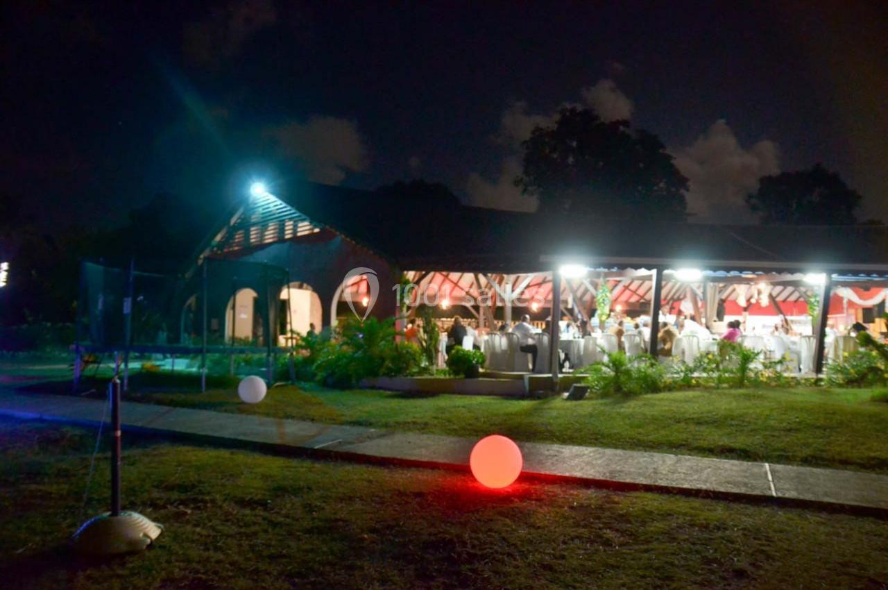 Salle éclairée en soirée avec des invités visibles à l'intérieur, entourée d'un jardin et de lumières décoratives.