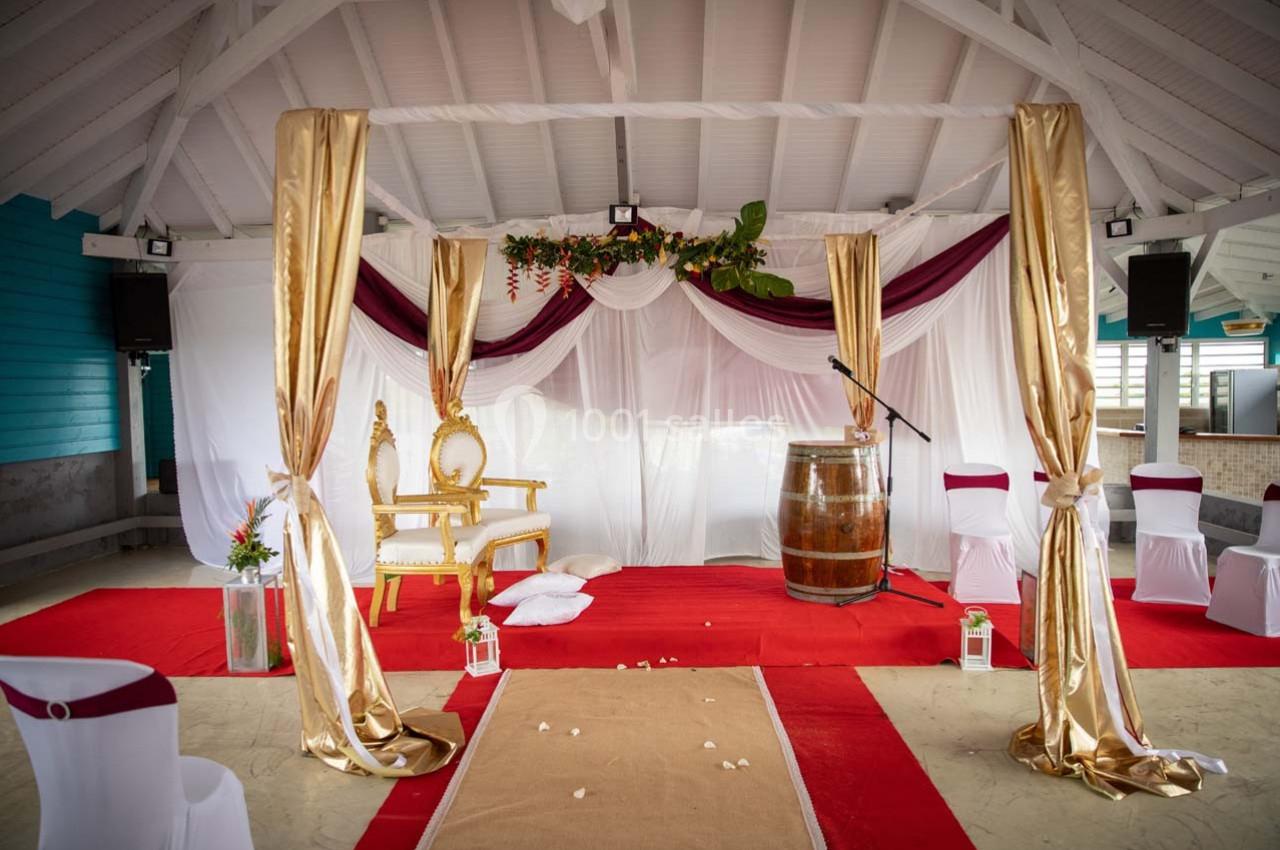 Décoration de cérémonie avec fauteuil doré, rideaux blancs et dorés, tapis rouge, tonneau en bois et chaises blanches.