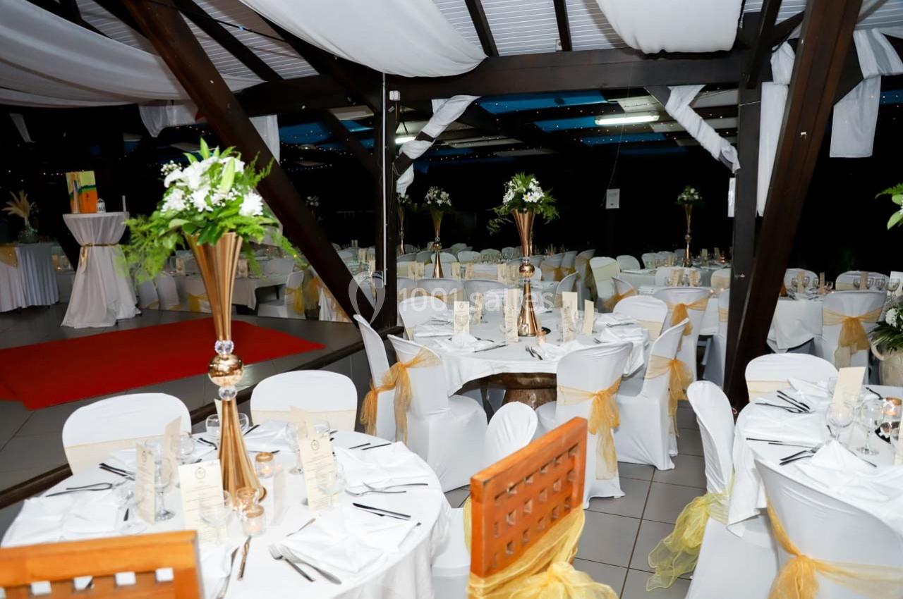 Salle de réception décorée avec des tables rondes, nappes blanches, chaises ornées de rubans jaunes et centres de table…