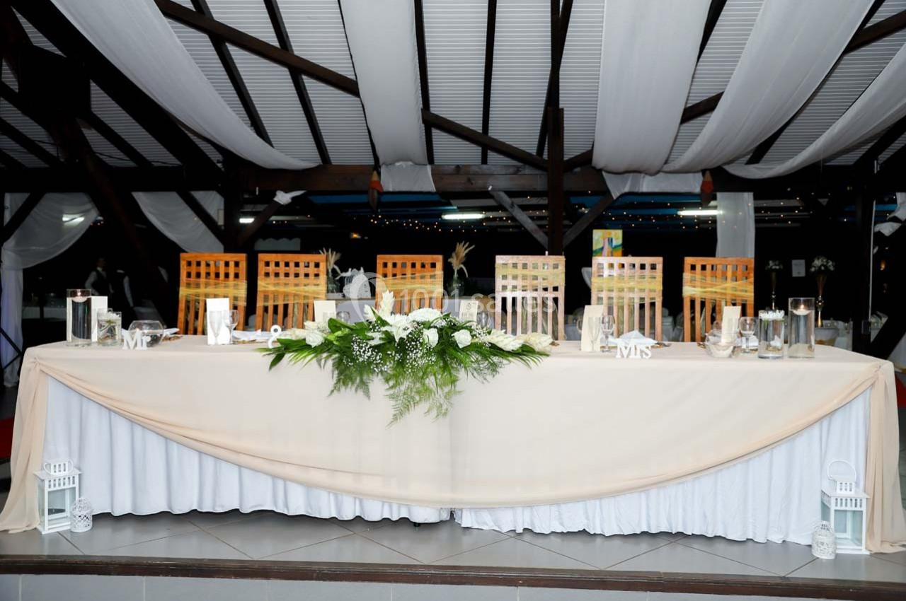 Grande table de réception décorée de nappes blanches, de lanternes, de fleurs et de chaises en bois sous un toit avec drapés.