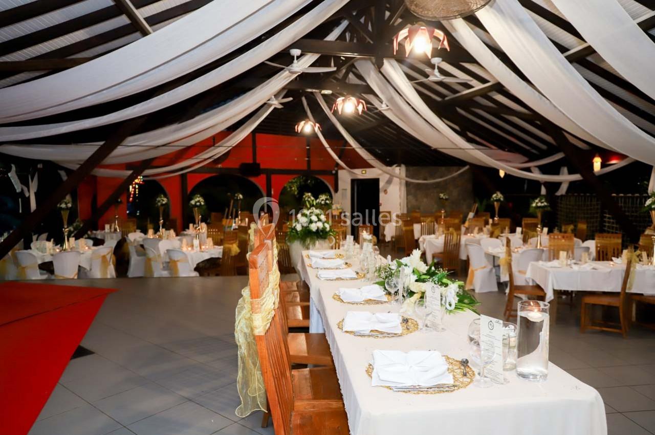 Salle de réception décorée avec des tables dressées, des chaises en bois et des drapés blancs suspendus au plafond.