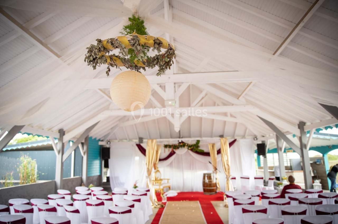 Salle décorée pour une cérémonie, avec chaises blanches, allée centrale rouge et arche ornée de tissus et feuillages.