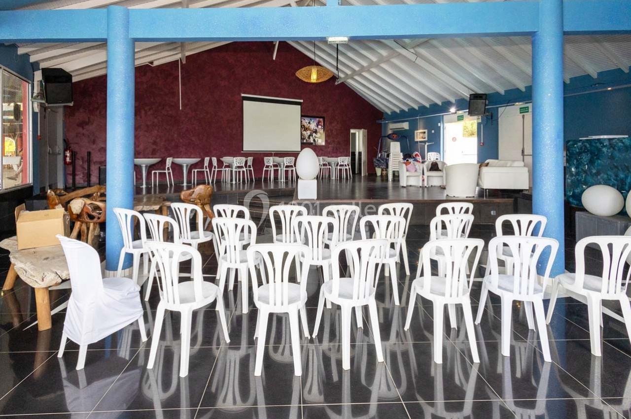 Salle avec des chaises blanches alignées face à un écran de projection, dans un espace lumineux avec un plafond en pente.