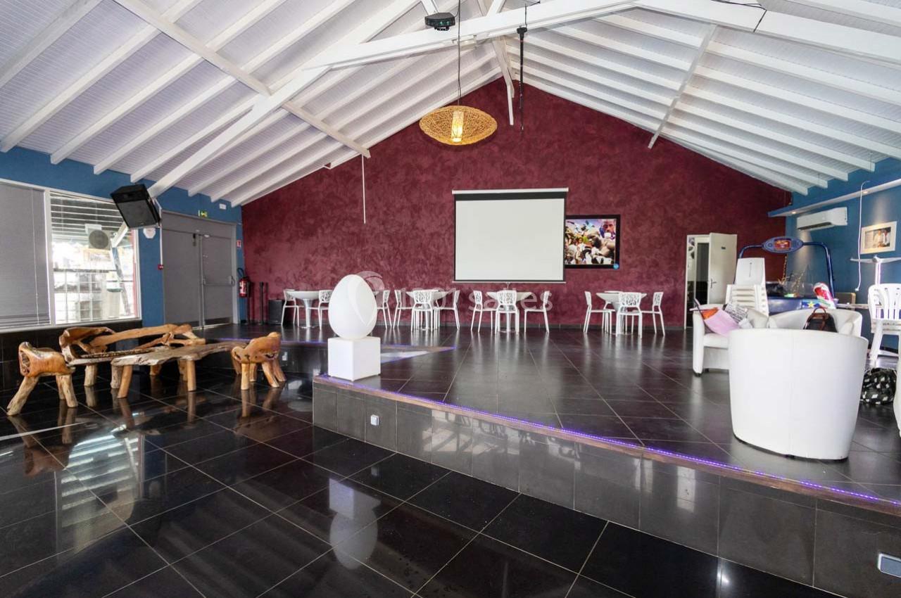 Salle spacieuse avec plafond en poutres blanches, murs rouges, tables et chaises blanches, et un écran de projection.