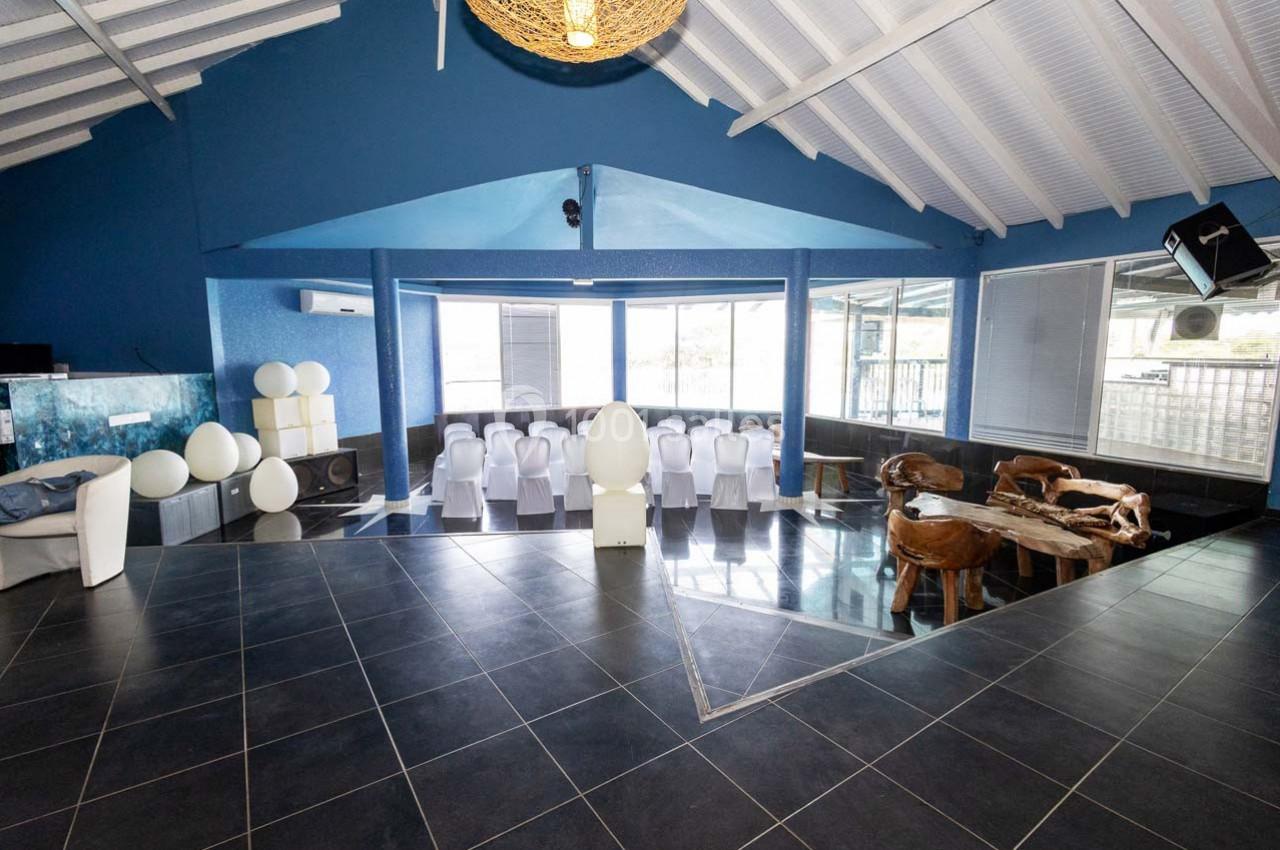 Salle lumineuse avec toit en pente, chaises blanches alignées, table en bois sculpté et grandes fenêtres donnant sur l…