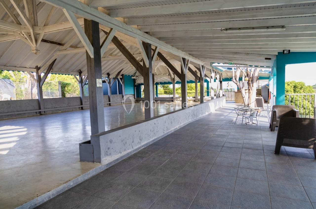 Grande terrasse couverte avec structure en bois, sol carrelé et quelques tables et chaises disposées près des ouvertures.