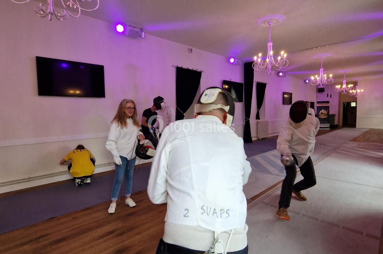 Deux personnes s'affrontent en escrime dans une salle éclairée, sous le regard de spectateurs en arrière-plan.