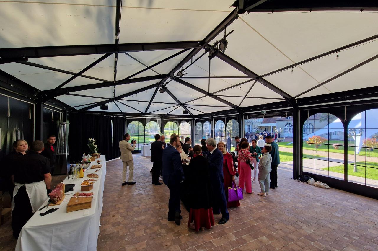 Groupe de personnes rassemblées dans une salle lumineuse avec buffet à gauche et vue sur un jardin à travers des baies…