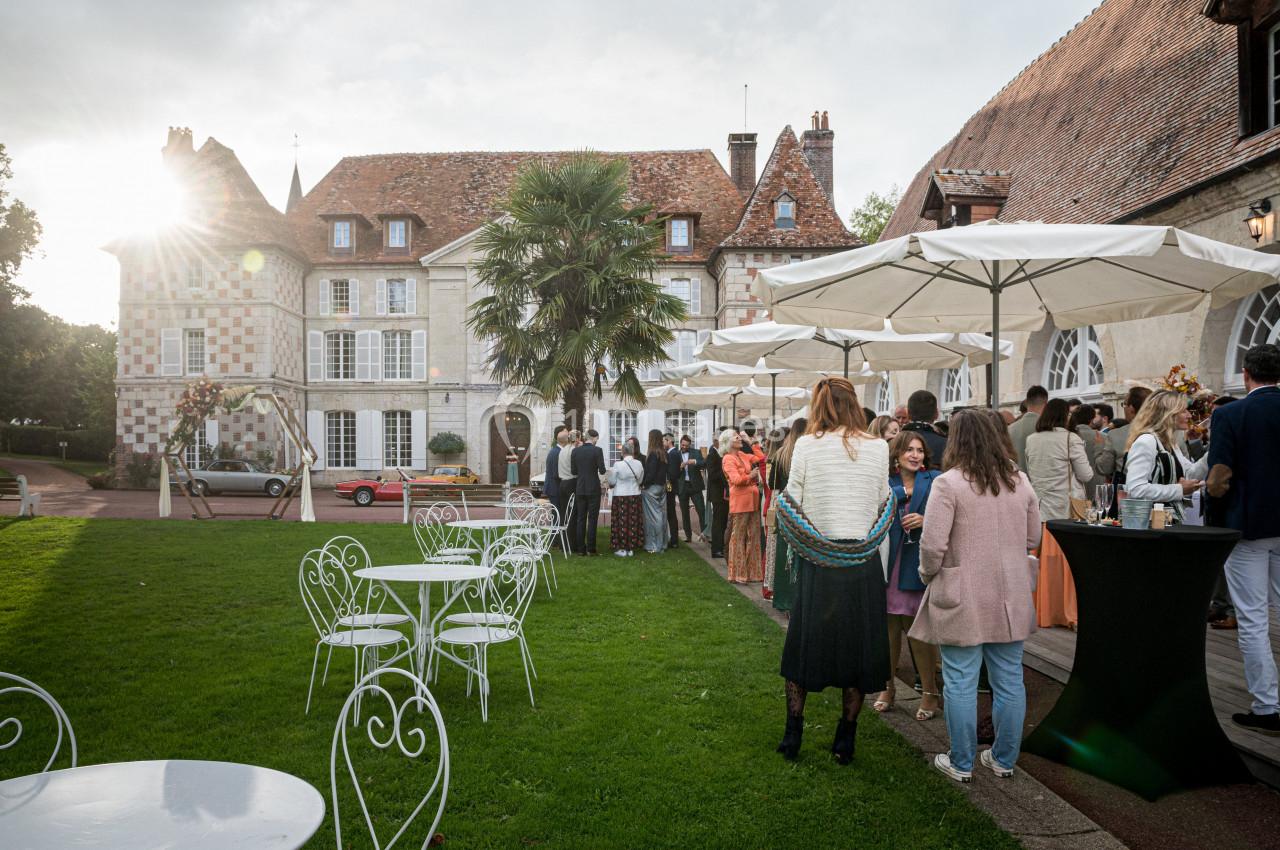 Groupe de personnes rassemblées dans le jardin d'un manoir, entourées de tables et parasols blancs.
