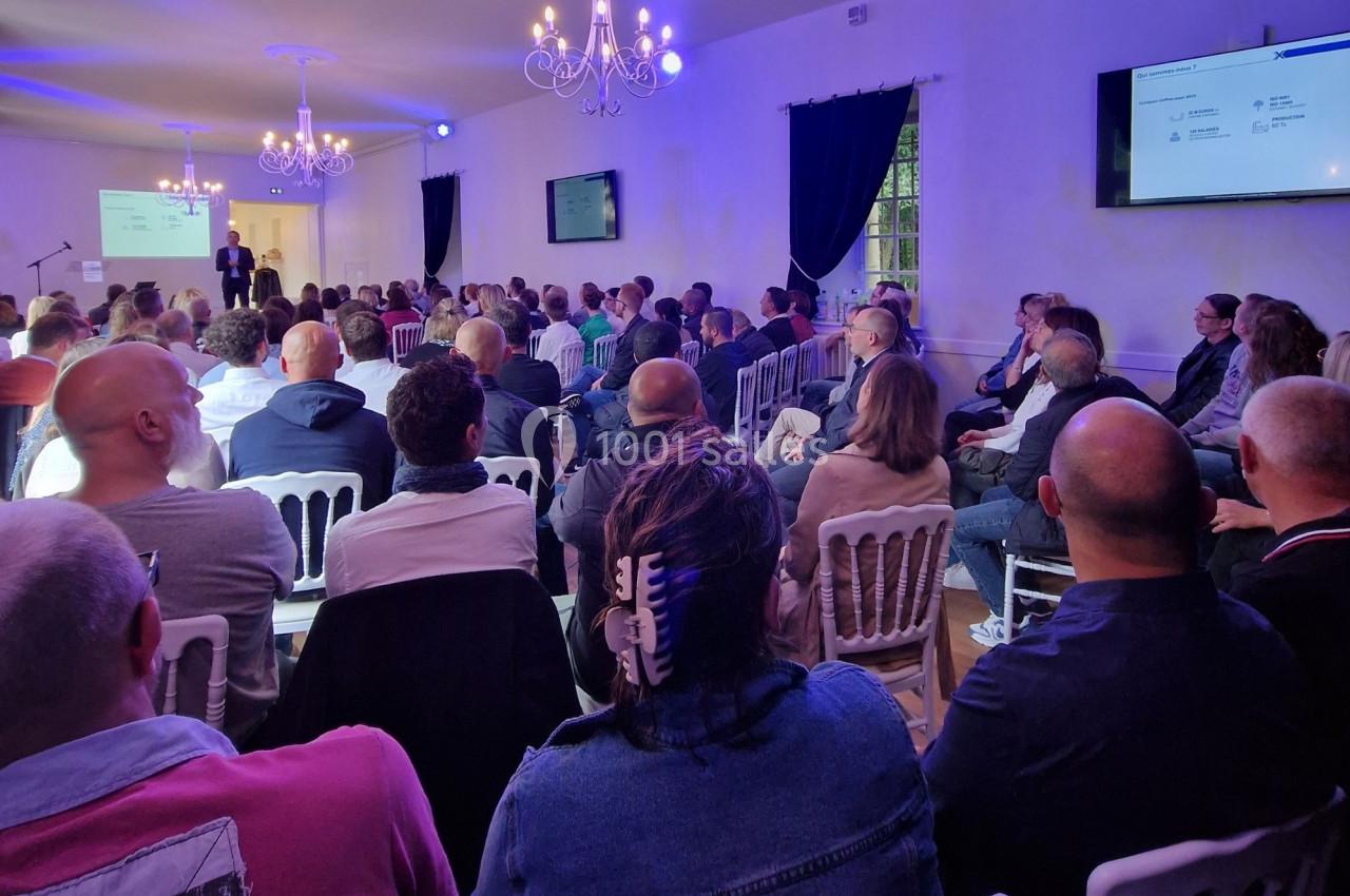 Un conférencier s'adresse à un public assis dans une salle éclairée, avec des écrans affichant des présentations.