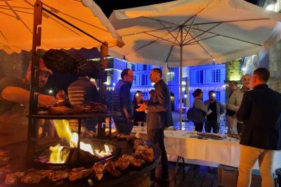 Des convives discutent près d'un barbecue en plein air, sous des parasols éclairés, dans une ambiance nocturne festive.