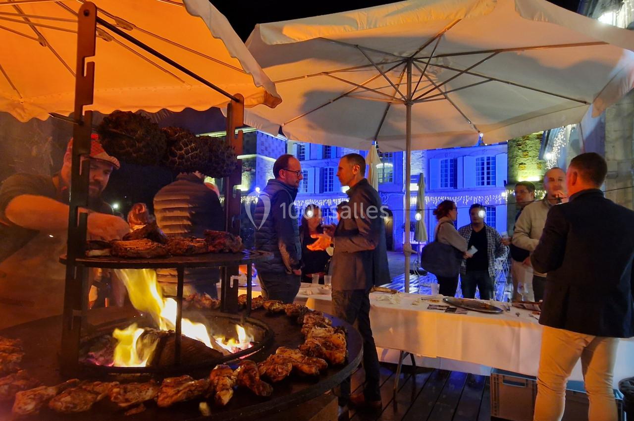 Des convives discutent près d'un barbecue en plein air, sous des parasols éclairés, dans une ambiance nocturne festive.