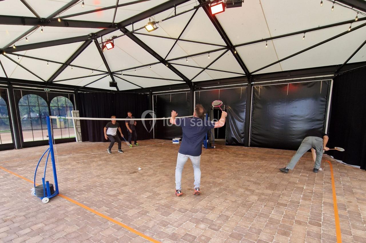 Quatre personnes jouent au badminton dans une structure couverte avec un sol en pavés et un filet central.