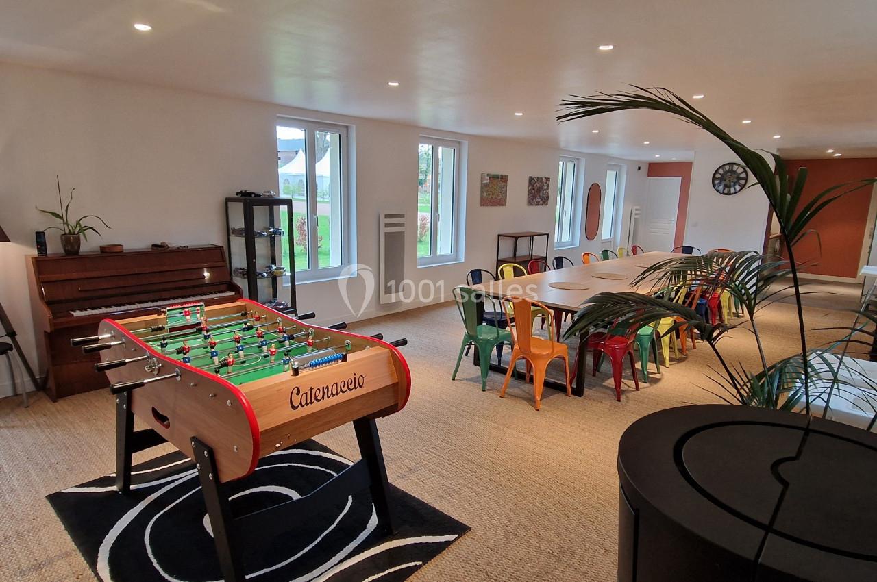 Salle lumineuse avec baby-foot, grande table entourée de chaises colorées, et vue sur un jardin à travers les fenêtres.