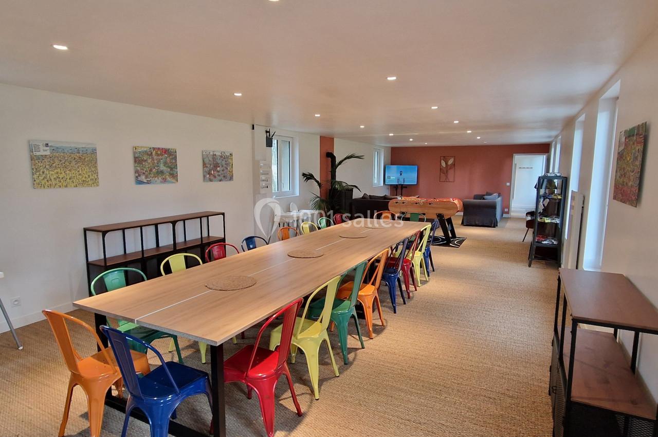 Salle lumineuse avec une grande table entourée de chaises colorées, des œuvres murales et un espace détente au fond.
