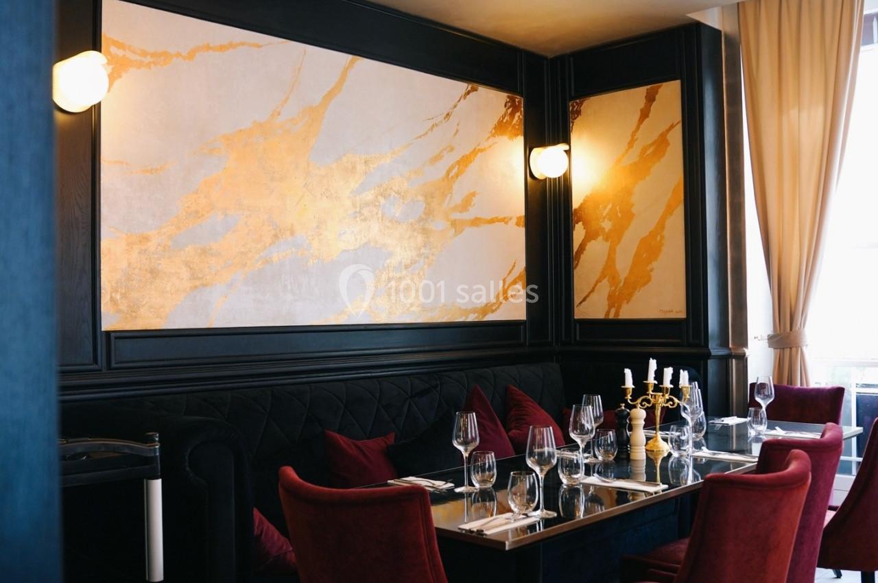 Salle de restaurant élégante avec banquettes noires, chaises rouges, table dressée et murs décorés de motifs dorés.