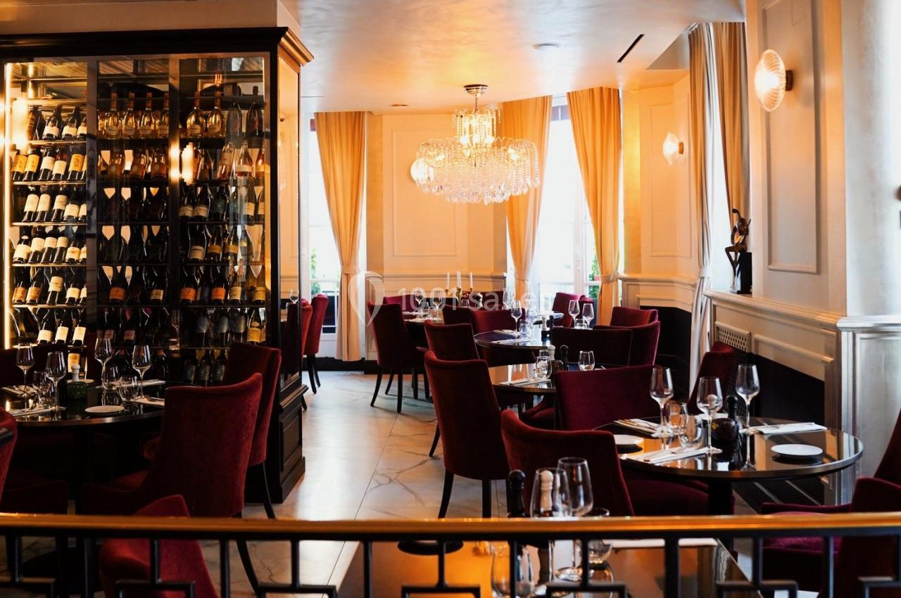 Salle de restaurant élégante avec chaises rouges, tables dressées et une cave à vin vitrée en arrière-plan.