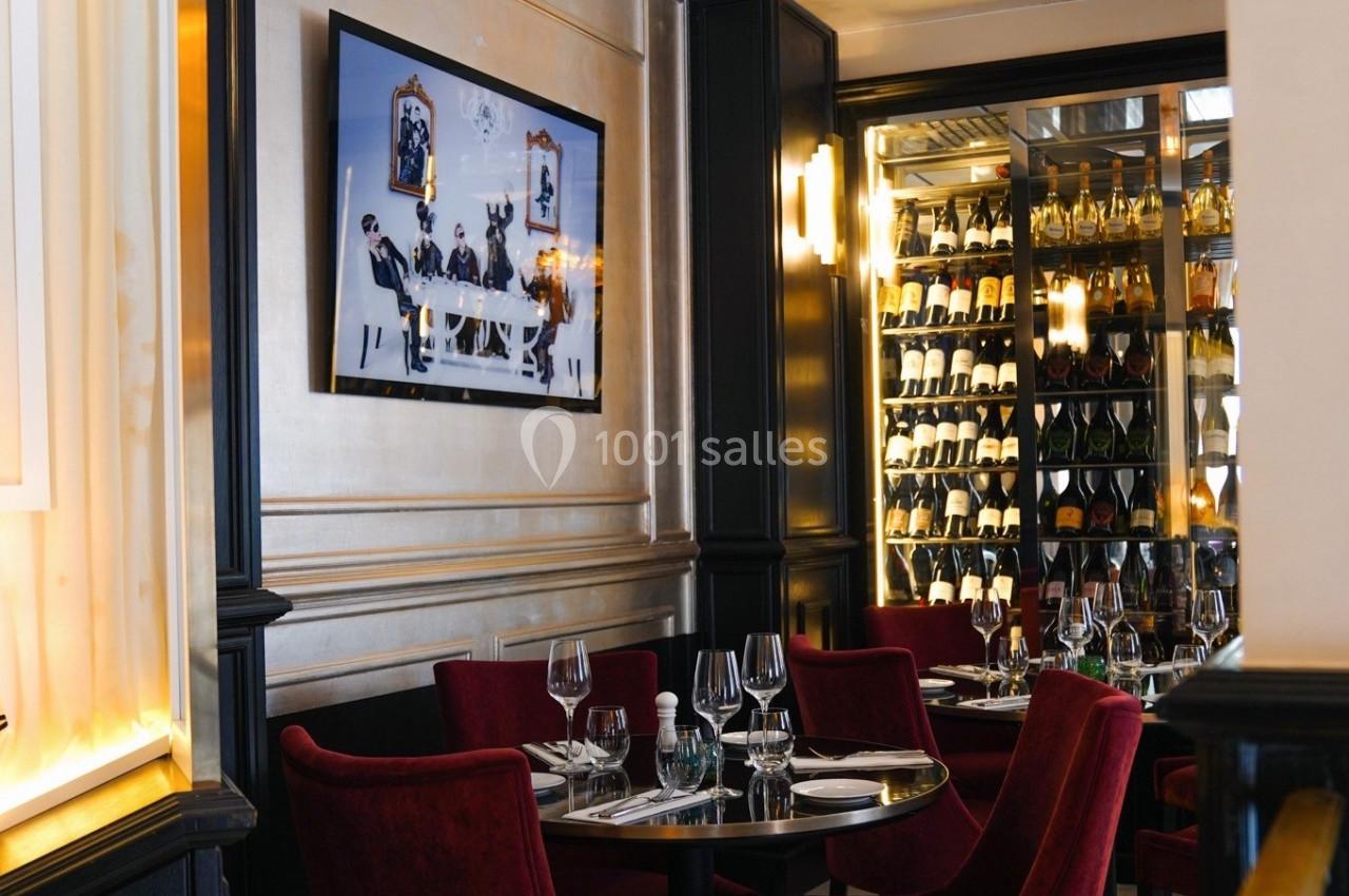 Salle de restaurant élégante avec tables dressées, chaises rouges, tableau mural et cave à vin vitrée en arrière-plan.