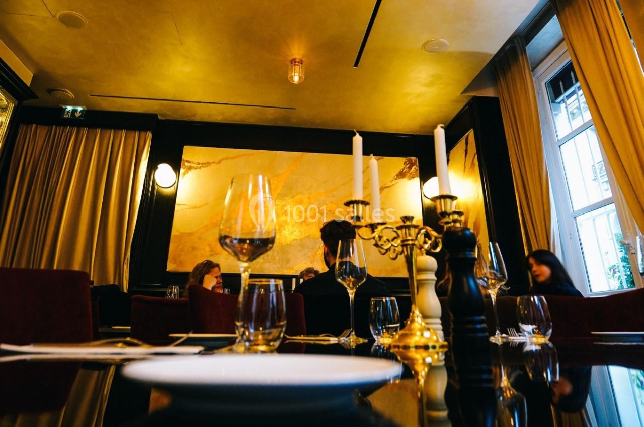 Salle de restaurant élégante avec des murs dorés, des chandeliers et des tables dressées avec des verres et assiettes.