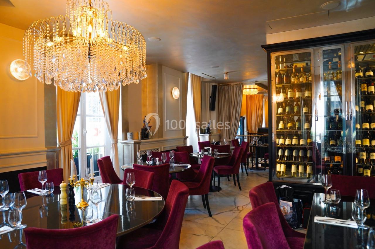 Salle de restaurant élégante avec lustre en cristal, chaises rouges, tables dressées et une cave à vin vitrée.
