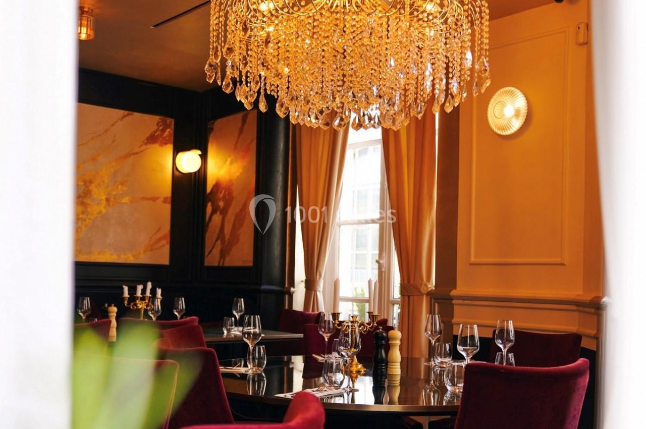 Salle de restaurant élégante avec lustre en cristal, rideaux beiges, murs sombres et tables dressées.