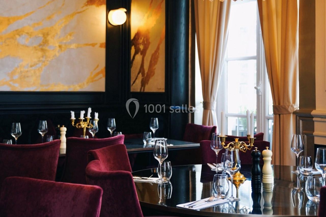 Salle de restaurant élégante avec chaises en velours rouge, tables dressées et lumière naturelle entrant par une fenêtre.