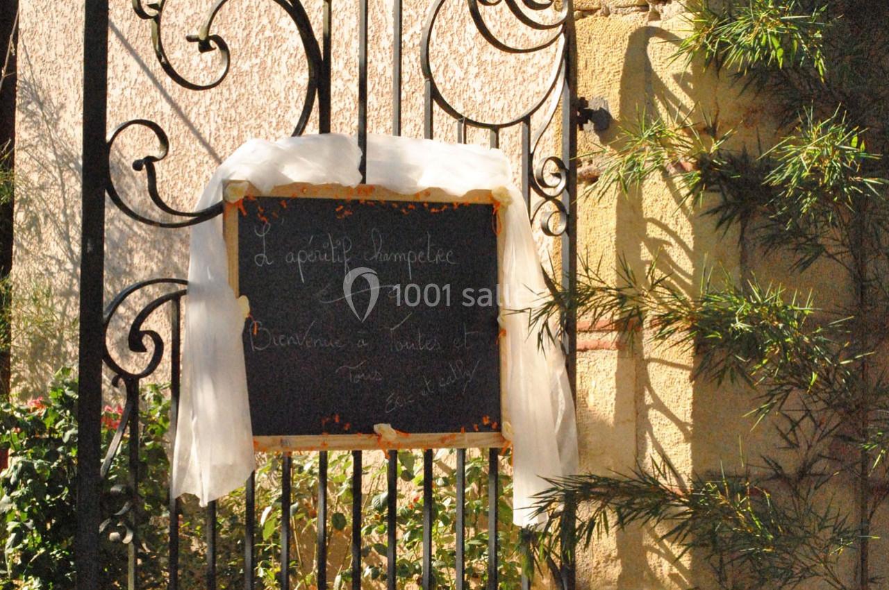 Ardoise décorative accrochée à une grille en fer forgé, entourée de végétation et d'un voile blanc.