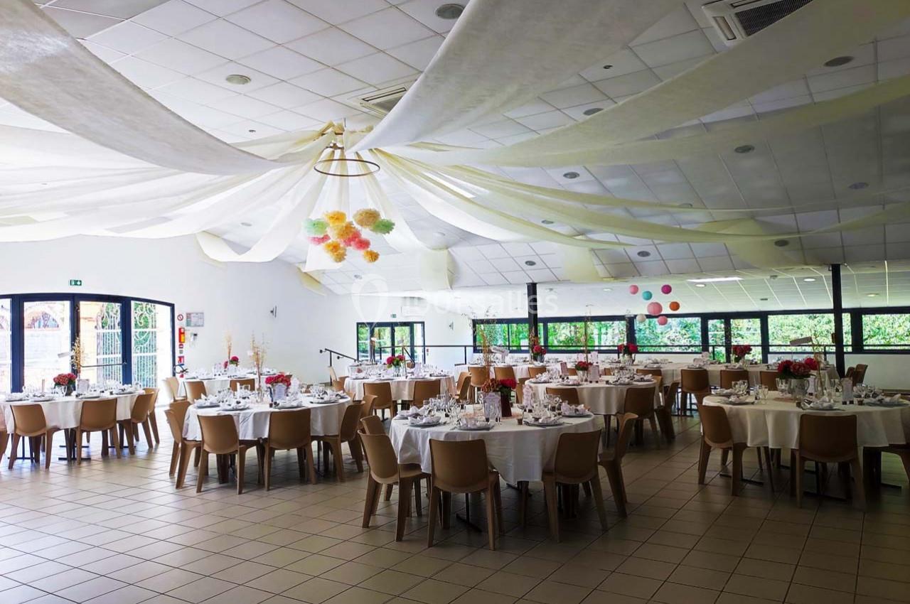 Salle de réception décorée avec des tables rondes dressées, des chaises en bois et des guirlandes suspendues au plafond.