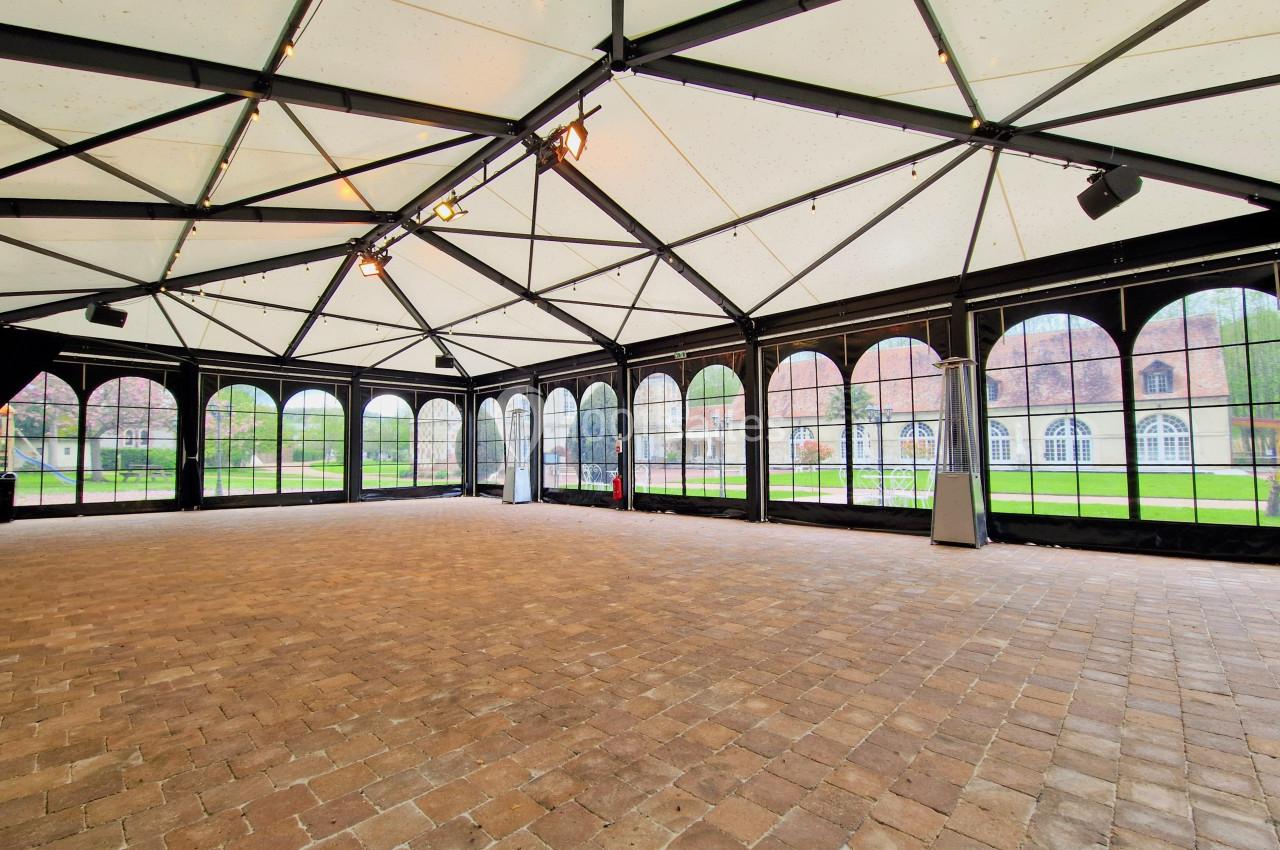 Intérieur d'une grande salle avec structure métallique, toit translucide et murs vitrés donnant sur un espace extérieur.