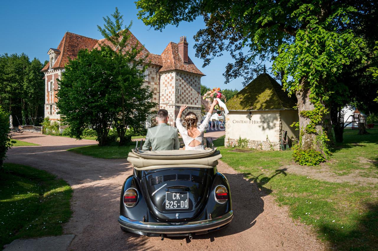 Un couple en tenue de mariage dans une voiture décapotable ancienne, devant un château entouré de verdure.