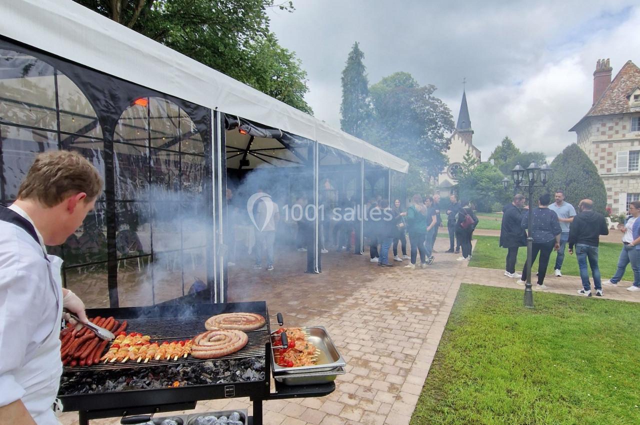Un cuisinier prépare un barbecue avec des saucisses et brochettes près d'une tente lors d'un événement en extérieur.