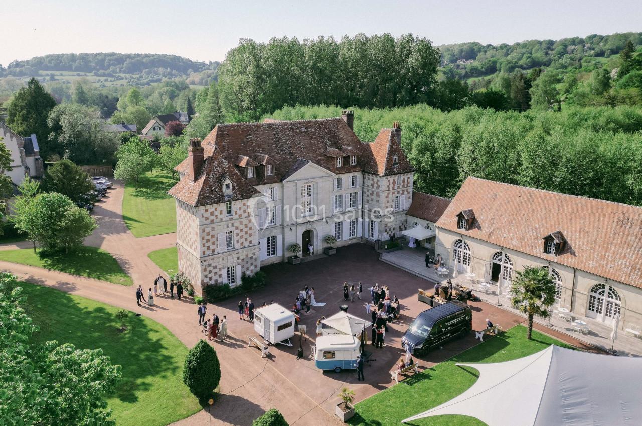 Vue aérienne d'un domaine avec un manoir en pierre, des dépendances, des arbres et des personnes rassemblées dans la cour.