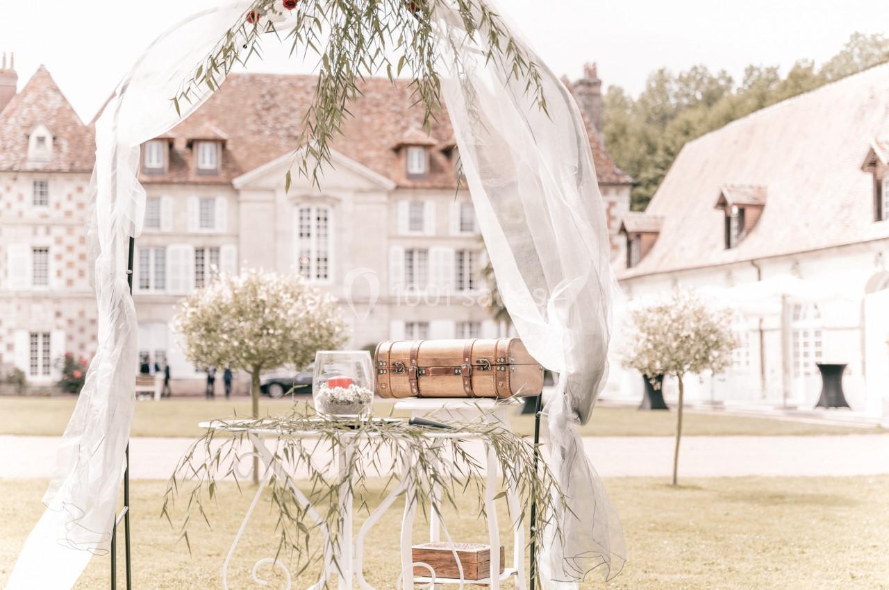 Arche décorée de voilages blancs et feuillages, avec une table ornée d'un coffre et d'une bougie, devant un manoir.