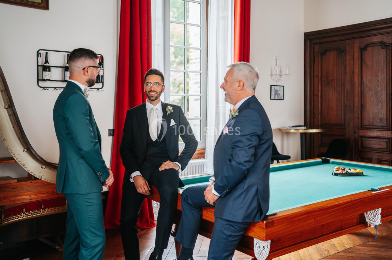 Trois hommes en costume discutent autour d'une table de billard dans une pièce élégante avec des rideaux rouges.
