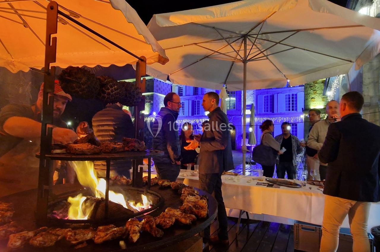Un chef cuisine des viandes sur un barbecue circulaire en plein air lors d'un événement nocturne sous des parasols éclairés.