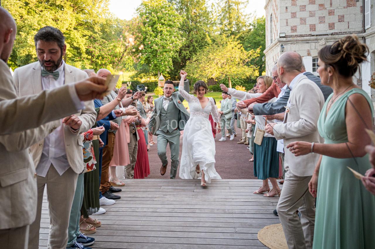 Un couple de mariés traverse une haie d'invités applaudissant sur une terrasse en bois, entourée de verdure.