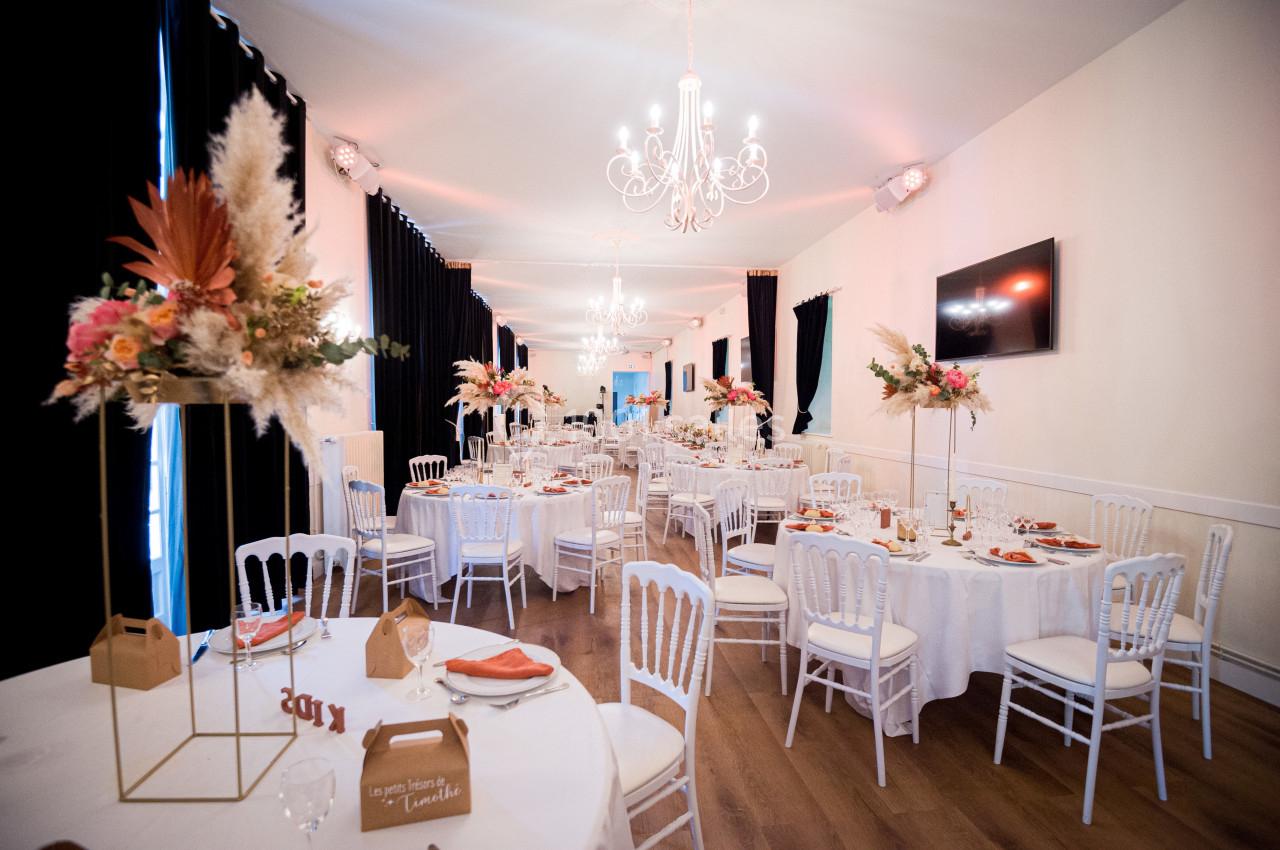 Salle de réception décorée avec des tables rondes, nappes blanches, chaises blanches et compositions florales élégantes.