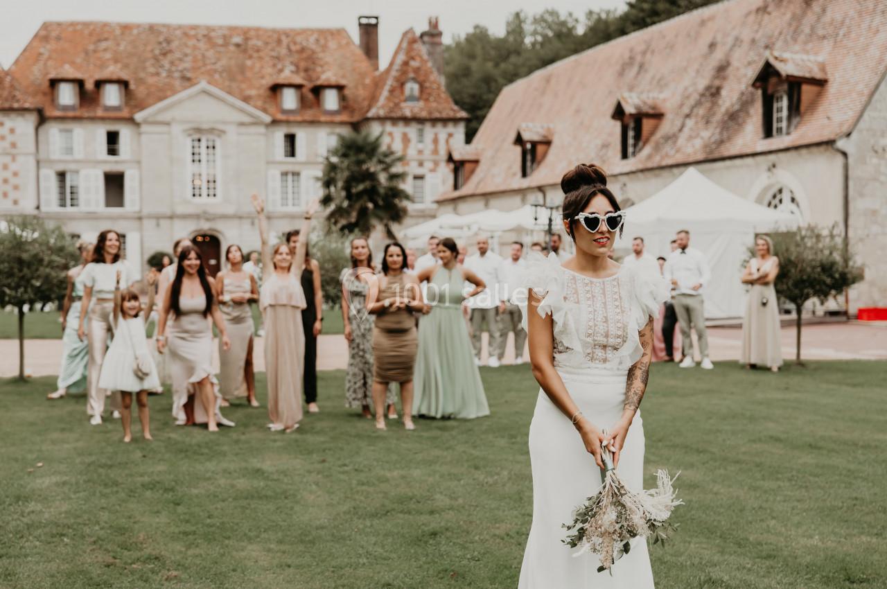 Une mariée tenant son bouquet se tient devant un groupe d'invités prêts à attraper le bouquet, dans un jardin près d'un…