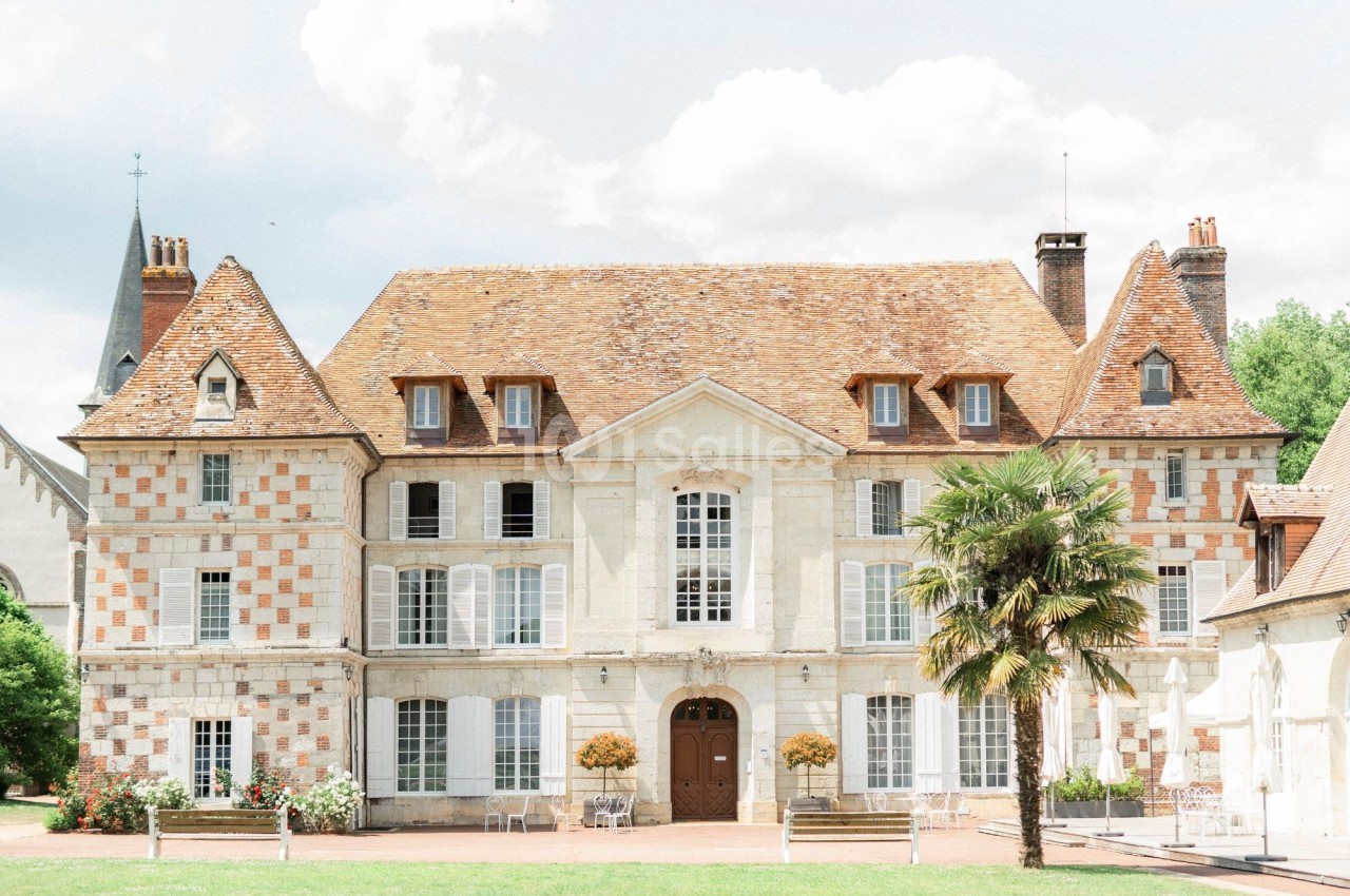 Façade d'un manoir en pierre et briques, avec toit en tuiles, entouré de verdure et d'une église en arrière-plan.