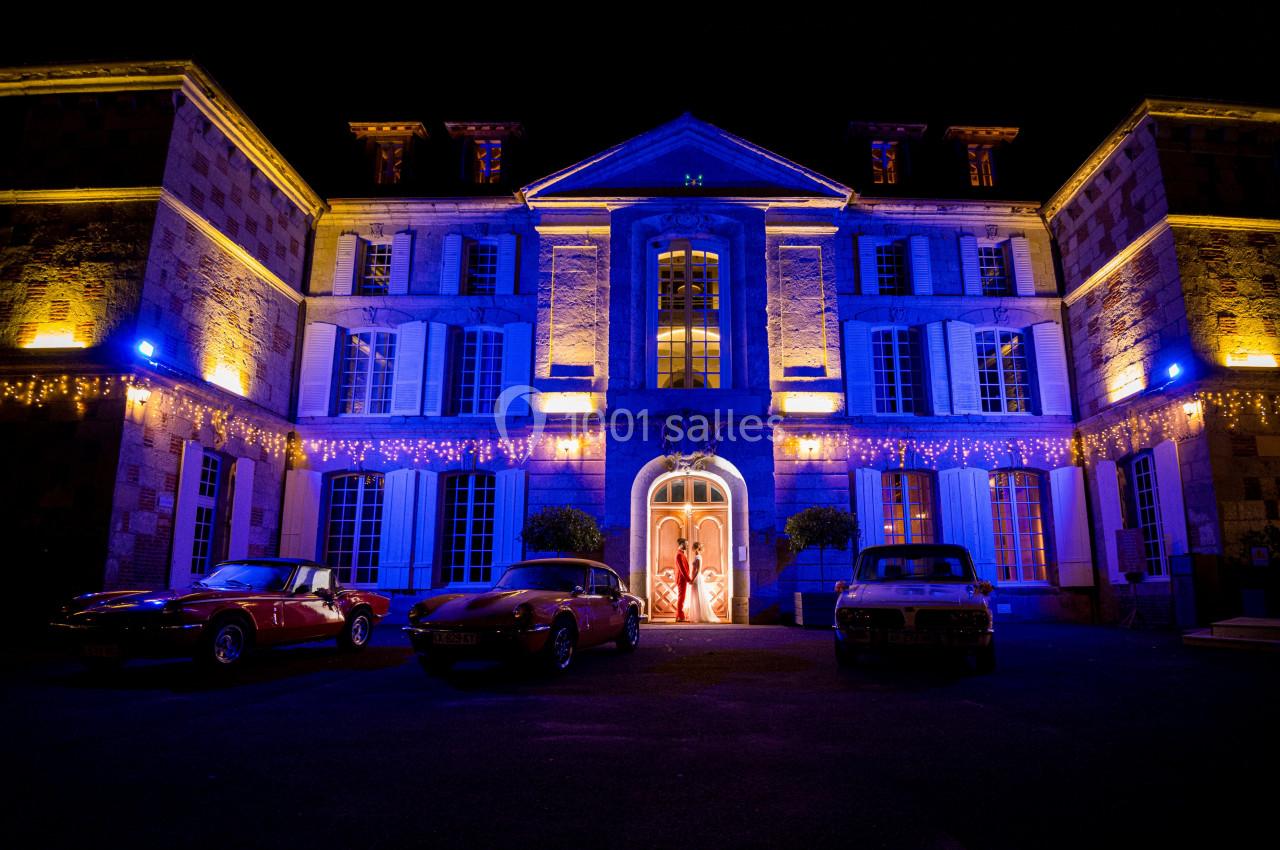 Façade d'un bâtiment illuminé en bleu et jaune la nuit, avec trois voitures anciennes stationnées devant.