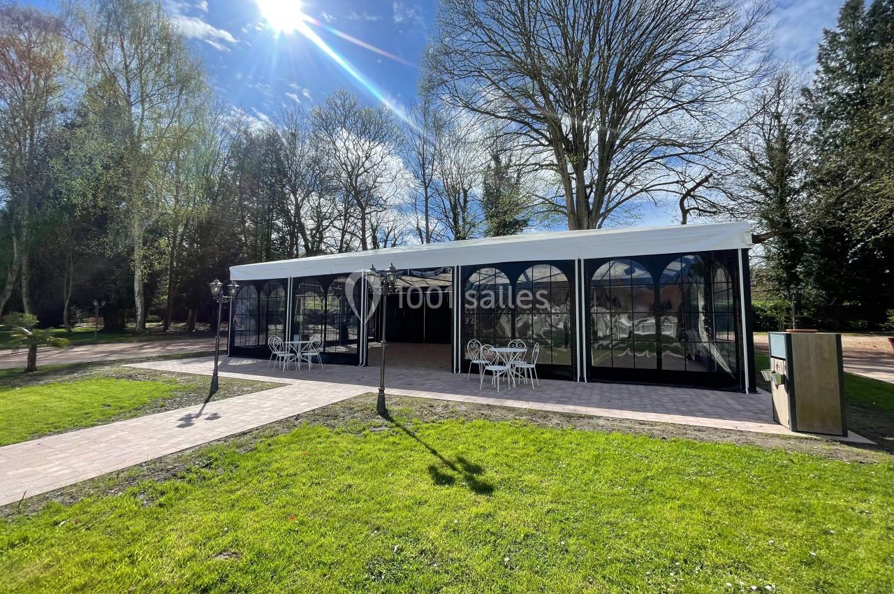 Structure extérieure avec grandes baies vitrées, entourée d'arbres, sous un ciel ensoleillé avec mobilier de jardin.