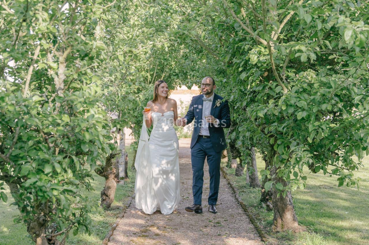 Un couple en tenue de mariage marche sur un sentier bordé d'arbres dans un jardin verdoyant.