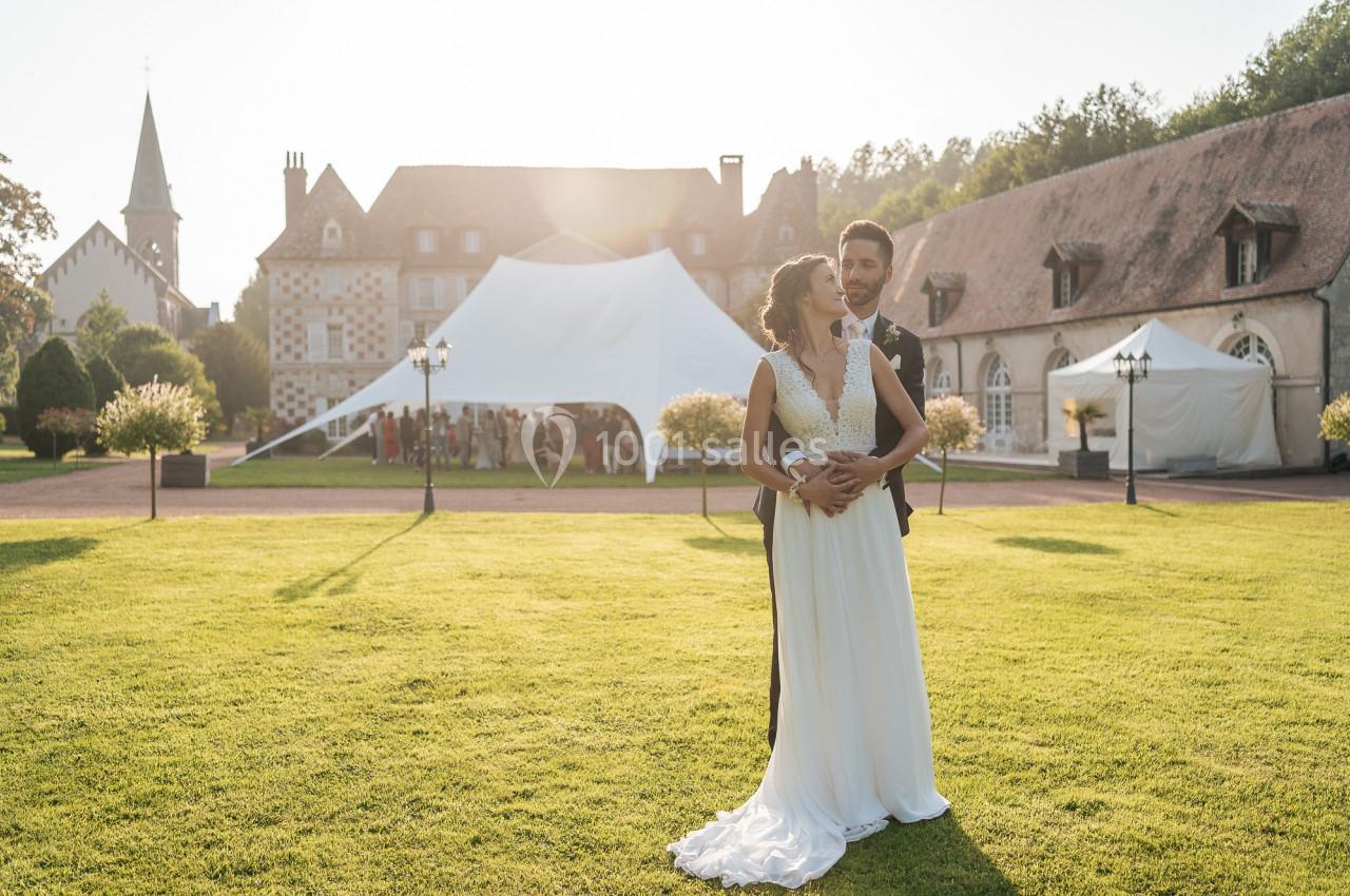 Un couple en tenue de mariage pose dans un jardin devant une tente blanche et des bâtiments anciens au coucher du soleil.