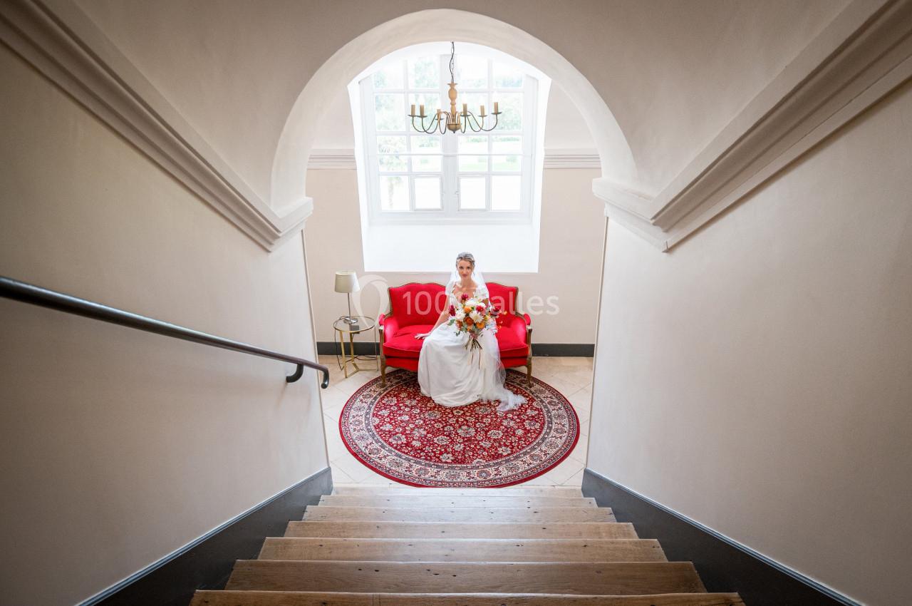 Une mariée en robe blanche est assise sur un canapé rouge, dans une pièce lumineuse avec un tapis rond.