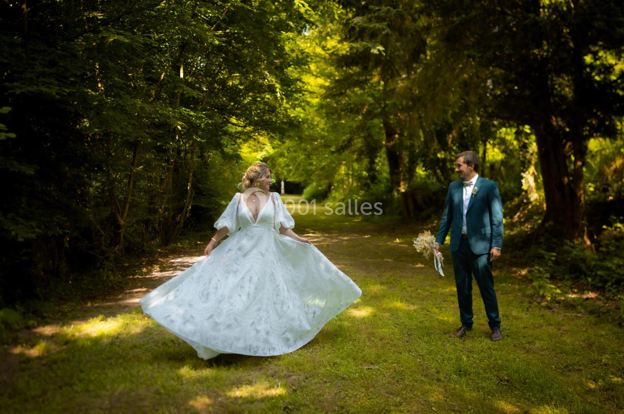 Une mariée en robe blanche tourne sur un chemin forestier, observée par un homme en costume bleu tenant un bouquet.