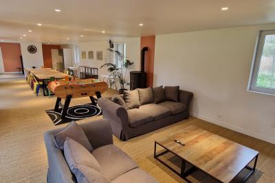 Salle spacieuse avec canapés, baby-foot, table de jeux et grandes fenêtres donnant sur un extérieur arboré.