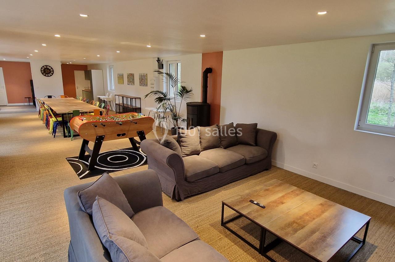 Salle spacieuse avec canapés, baby-foot, table de jeux et grandes fenêtres donnant sur un extérieur arboré.