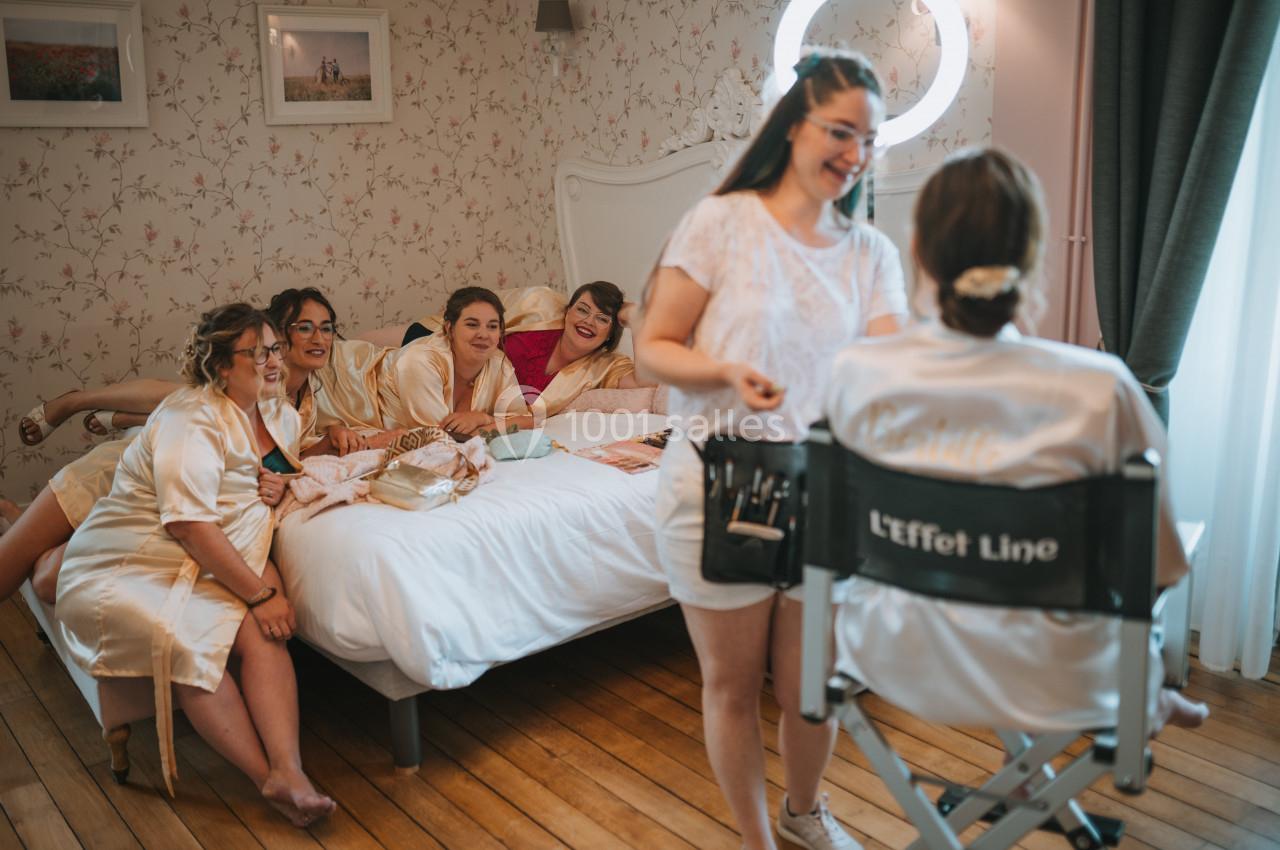 Des femmes en robes de satin rient et discutent sur un lit pendant qu'une maquilleuse prépare une mariée assise.